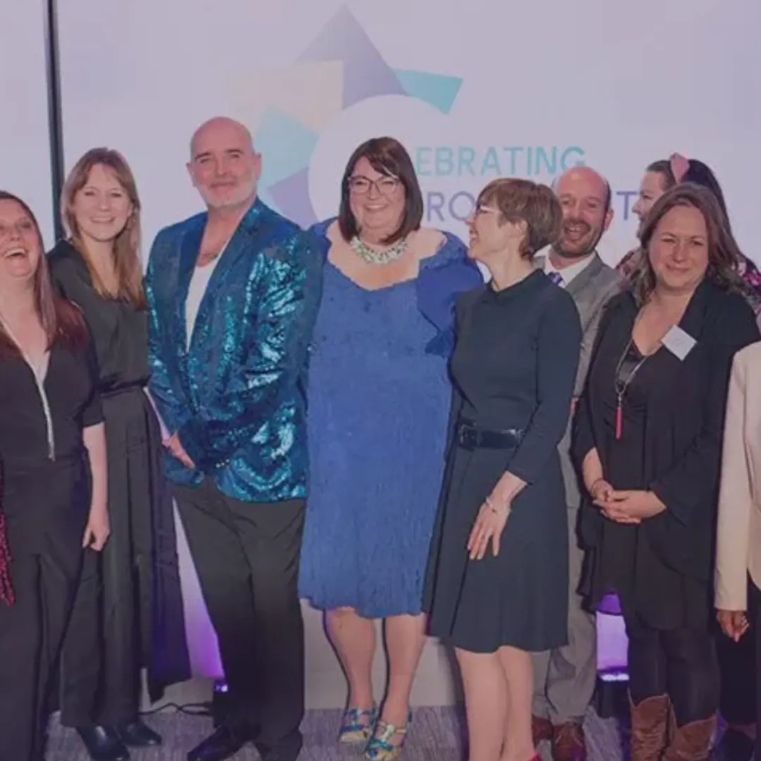 A woman with short brown hair and glasses, wearing a blue dress and smiling whilst surrounded by colleagues in a professional setting.