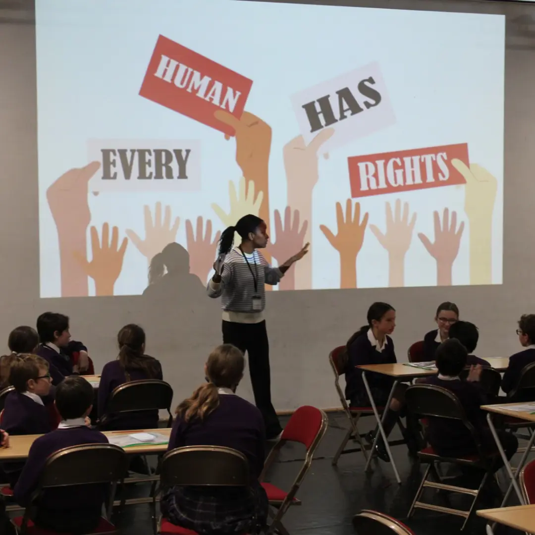 A Black woman with her hair in a ponytail presents to a classroom of secondary school students wearing purple uniforms. She stands in front of a large projected slide displaying "EVERY HUMAN HAS RIGHTS" with illustrated diverse hands raised up. The students sit at individual desks arranged in rows, attentively listening to the human rights presentation in what appears to be a school assembly hall or classroom.