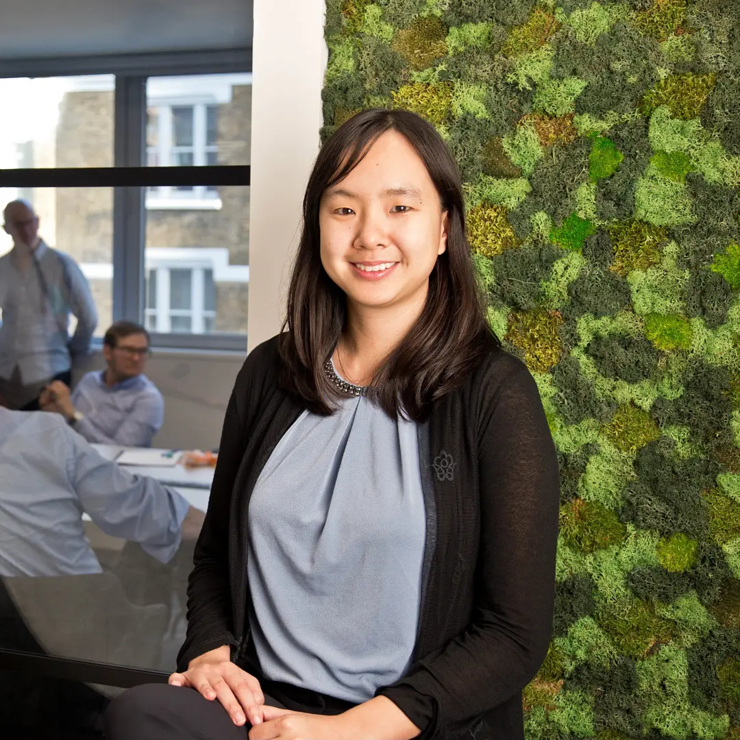 A woman of Asian heritage with shoulder-length dark hair, smiling and wearing a grey top with a black cardigan. She stands in a modern office space with a decorative moss wall behind her and colleagues visible in the background.