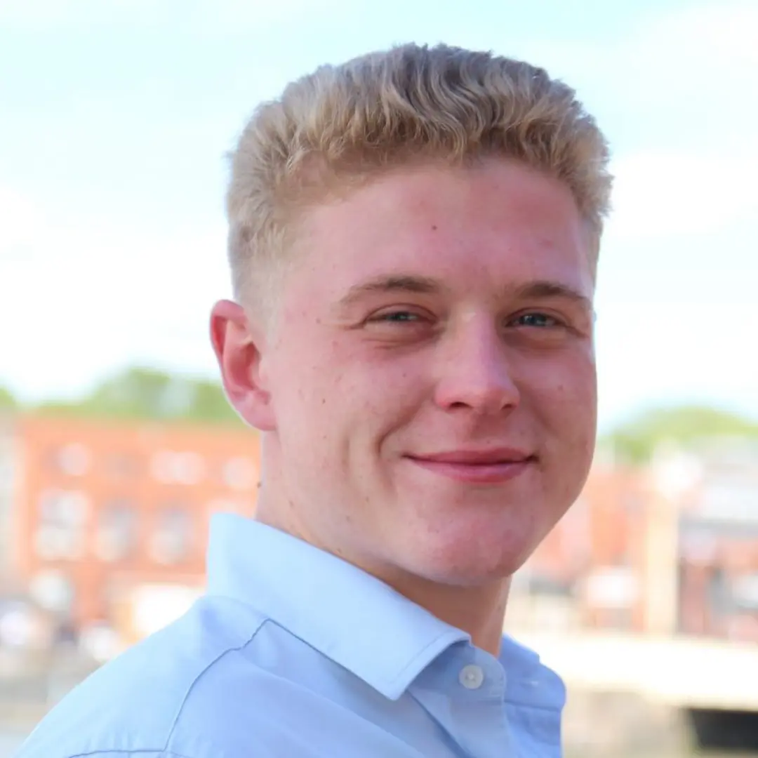 Young white man with short blond hair wearing a light blue collared shirt, smiling slightly while standing outdoors in front of a blurred background of red-brick buildings and a bright sky.