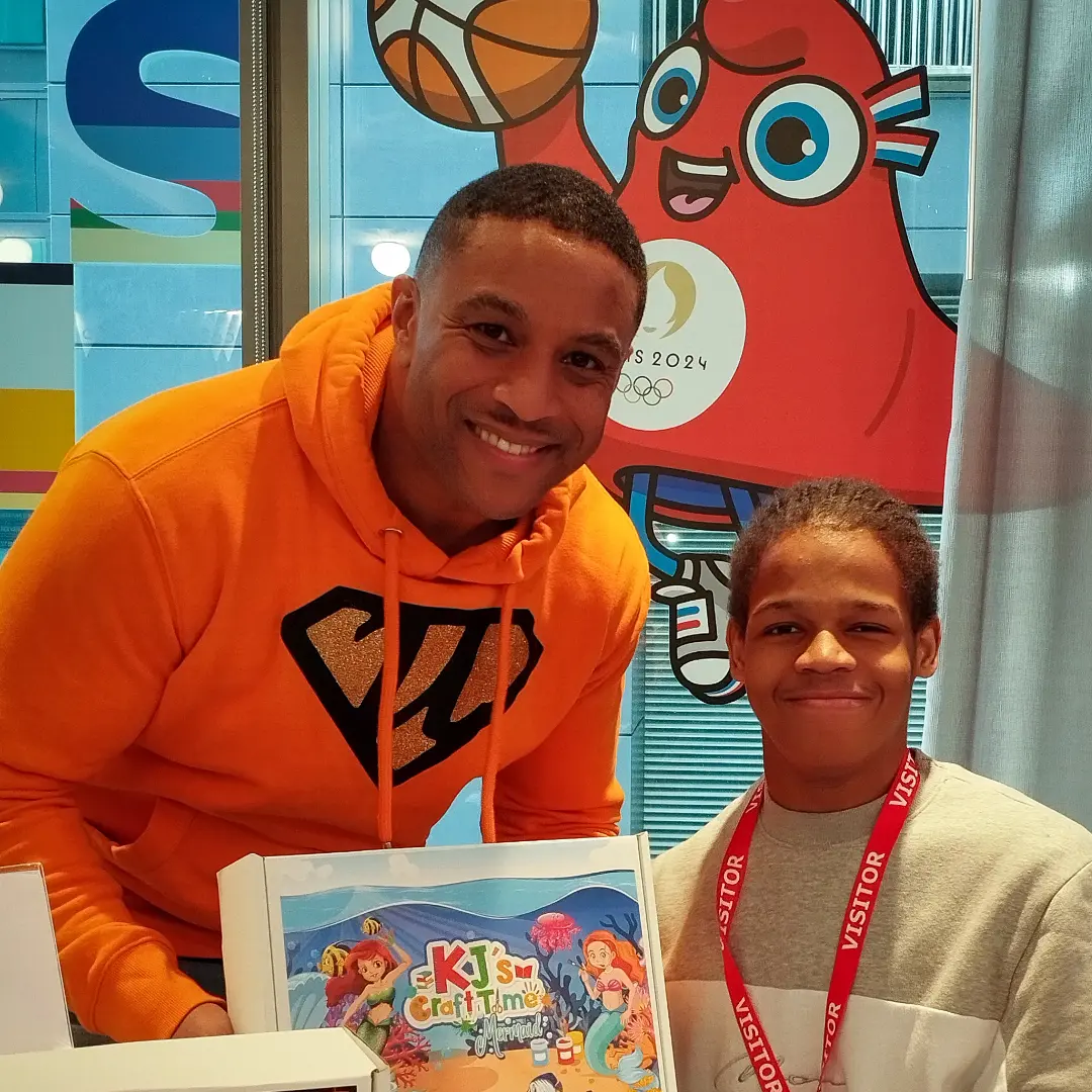 A Black man in an orange hoodie, smiling as he crouches down beside a boy of approximately ten years old for the photograph.