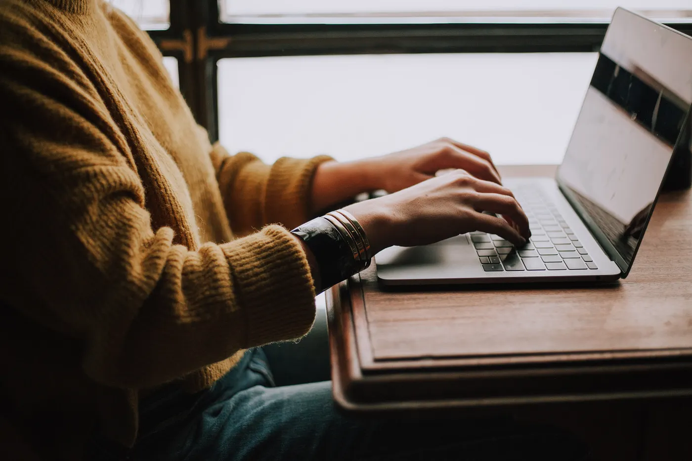 A person sits at an open laptop wearing a mustard coloured jumper