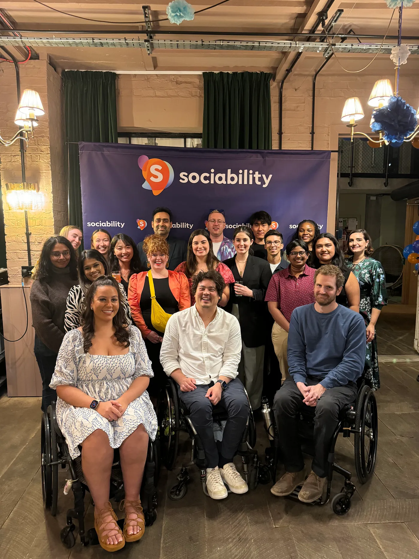 Matt, CEO and Founder of Sociability is a man in his 30s in a white shirt sitting on a wheelchair. He is smiling and surrounded by people, there is a purple banner behind them with Sociability written across it.